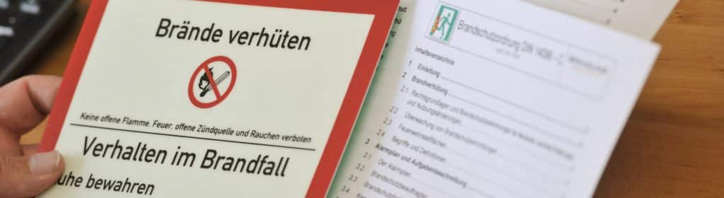 A person is holding a German safety brochure entitled "Brände verhüten" (Preventing fires) with a no-smoking symbol, next to a document in which fire protection windows and fire protection doors from Brandschutz Tischlerei Beelitz are listed in the table of contents