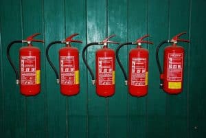 Fünf rote Feuerlöscher sind in einer Reihe an einer grünen Holzwand montiert, ideal für Umgebungen mit Brandschutzfenster oder einer Brandschutztür von Brandschutz Tischlerei Beelitz. Jeder Feuerlöscher hat einen Schlauch und weiße Aufkleber mit Anweisungen und Symbolen auf der Vorderseite.