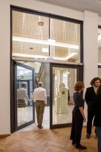 Mehrere Personen stehen und gehen durch eine große Brandschutztür mit einem Metallrahmen in einem modernen Gebäude. Der Innenraum ist mit Holzböden, hohen Decken und netzartigen Glasscheiben ausgestattet, die die Handwerkskunst der Brandschutz Tischlerei Beelitz demonstrieren.