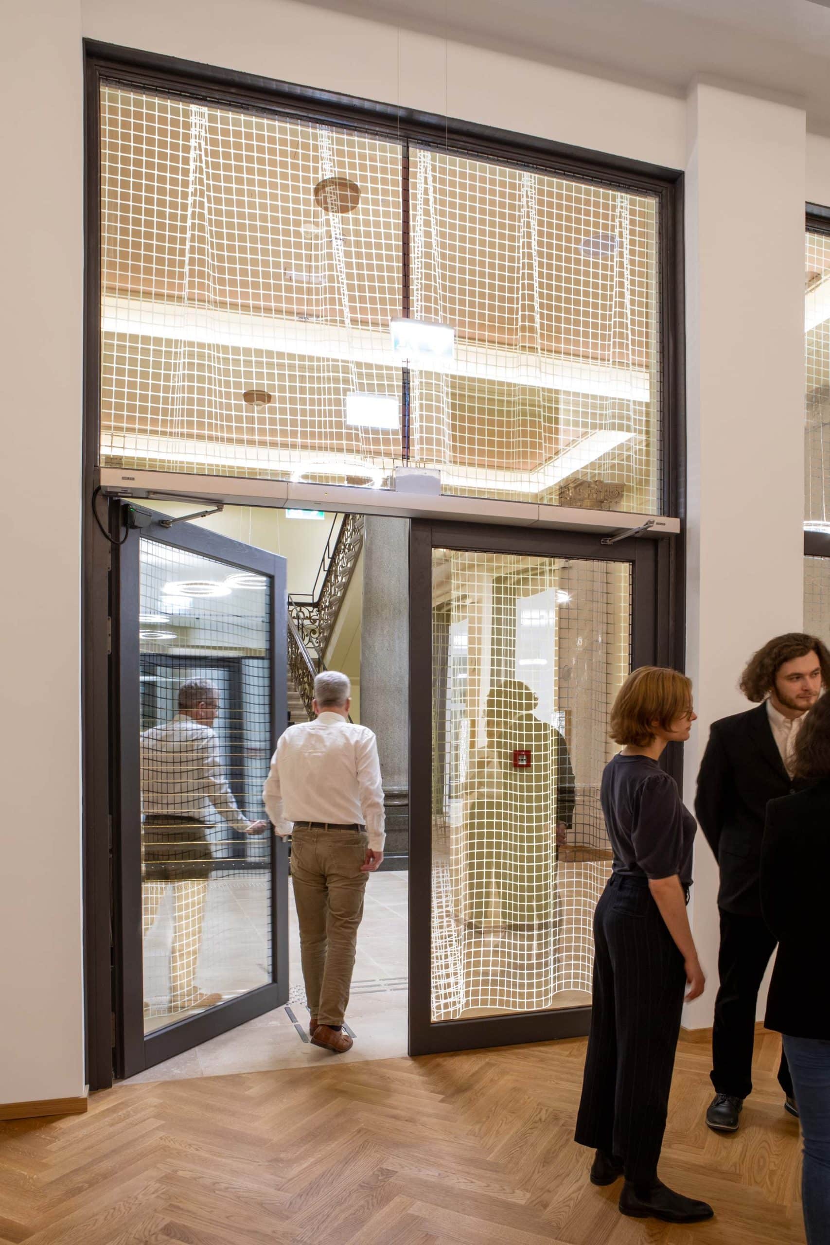 Mehrere Personen stehen und gehen durch eine große Brandschutztür mit einem Metallrahmen in einem modernen Gebäude. Der Innenraum ist mit Holzböden, hohen Decken und netzartigen Glasscheiben ausgestattet, die die Handwerkskunst der Brandschutz Tischlerei Beelitz demonstrieren.