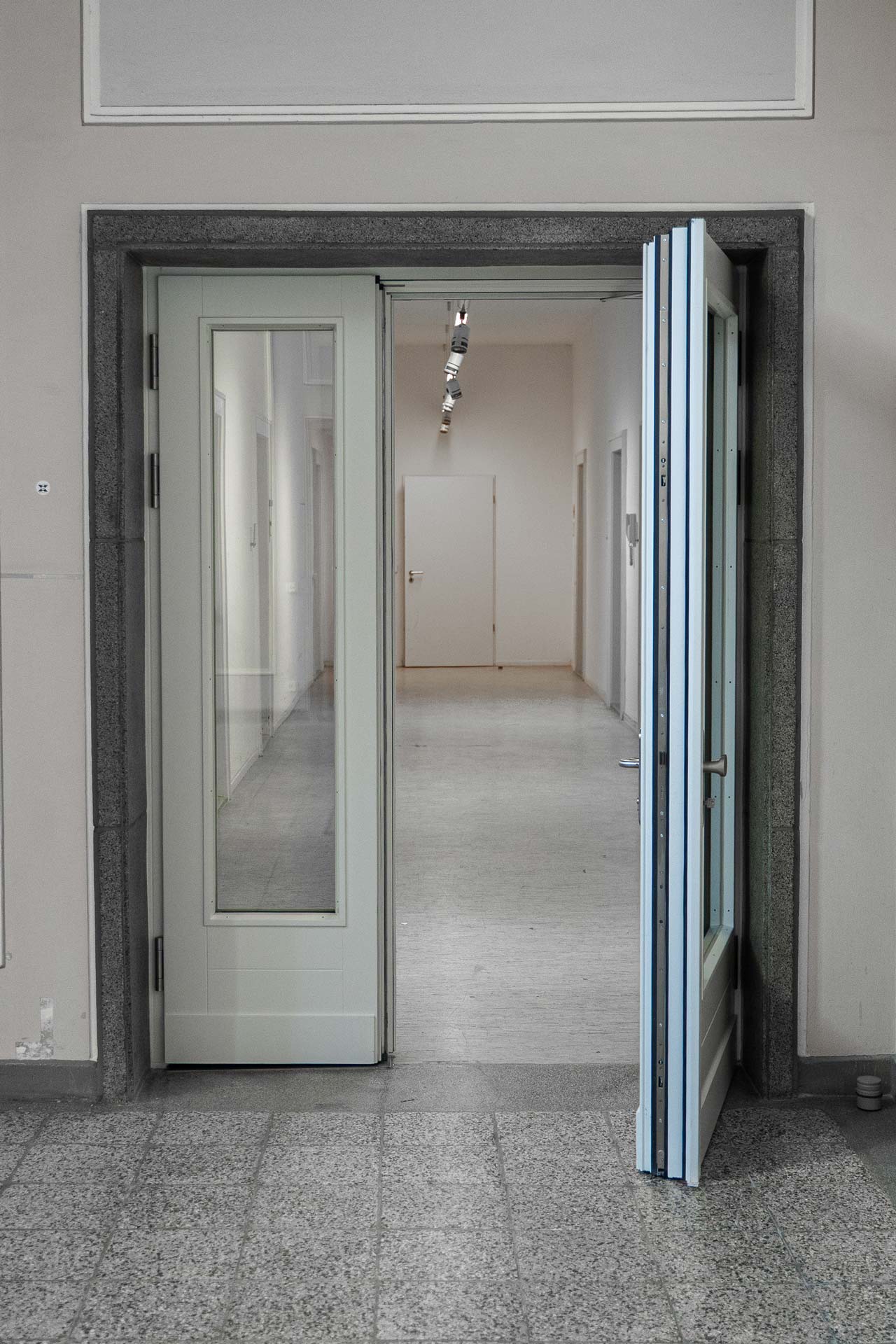 Eine Reihe von Doppeltüren, darunter eine spezielle Brandschutztür, von denen eine teilweise geöffnet ist, führt in einen gut beleuchteten Flur mit weißen Wänden, sichtbaren Deckenleuchten und mehreren geschlossenen Türen entlang des Korridors.