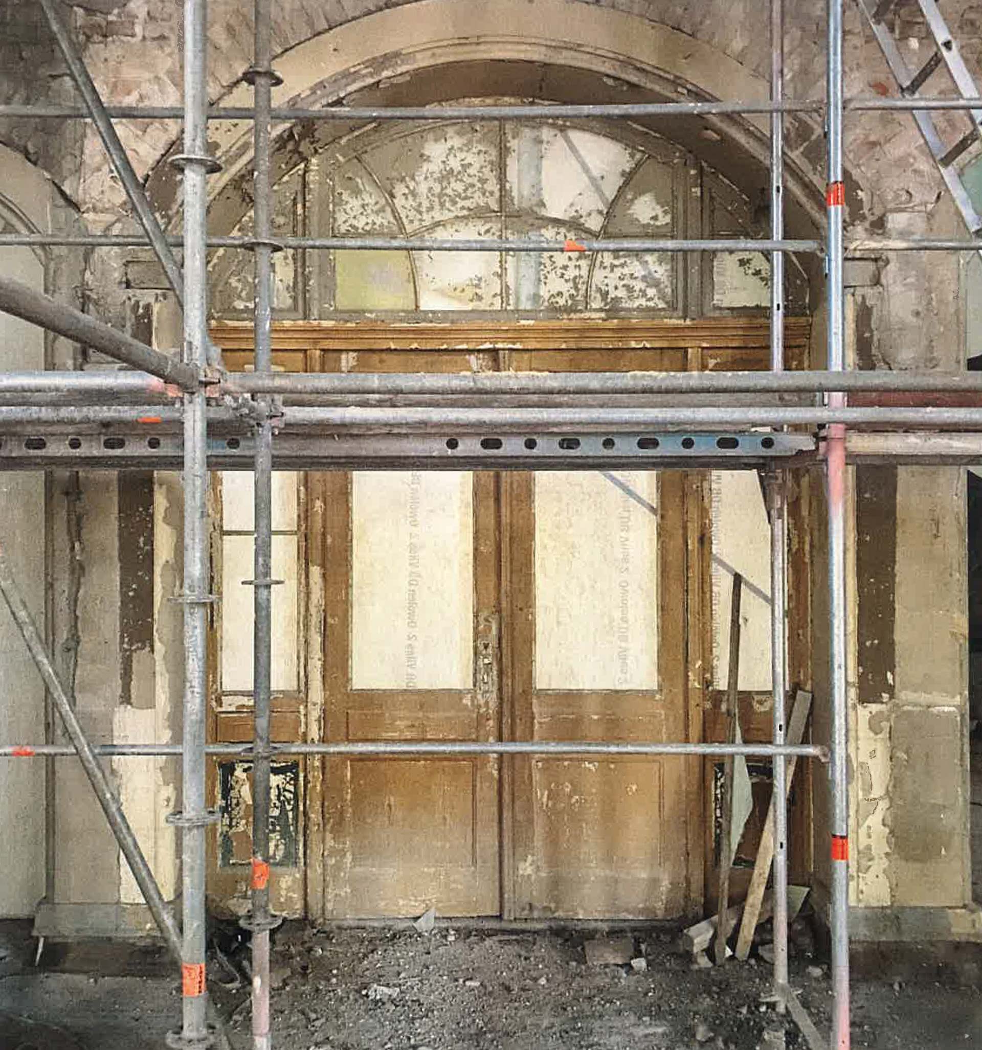 Ein Baugerüst steht vor einer großen, verwitterten Holztür mit darüber liegenden Rundbogenfenstern. Die umgebende Wand zeigt abblätternde Farbe und Verfallsspuren, was auf Renovierungsarbeiten der Brandschutz Tischlerei Beelitz für eine neue Brandschutztür oder ein Brandschutzfenster schließen lässt.