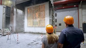Zwei Personen mit orangefarbenen Schutzhelmen beobachten, wie ein großes Brandschutzfenster auf seine Feuerbeständigkeit geprüft wird. Flammen und Rauch steigen im Prüfbereich auf, während die Sicherheitsausrüstung der Brandschutz Tischlerei Beelitz die Szene umgibt.