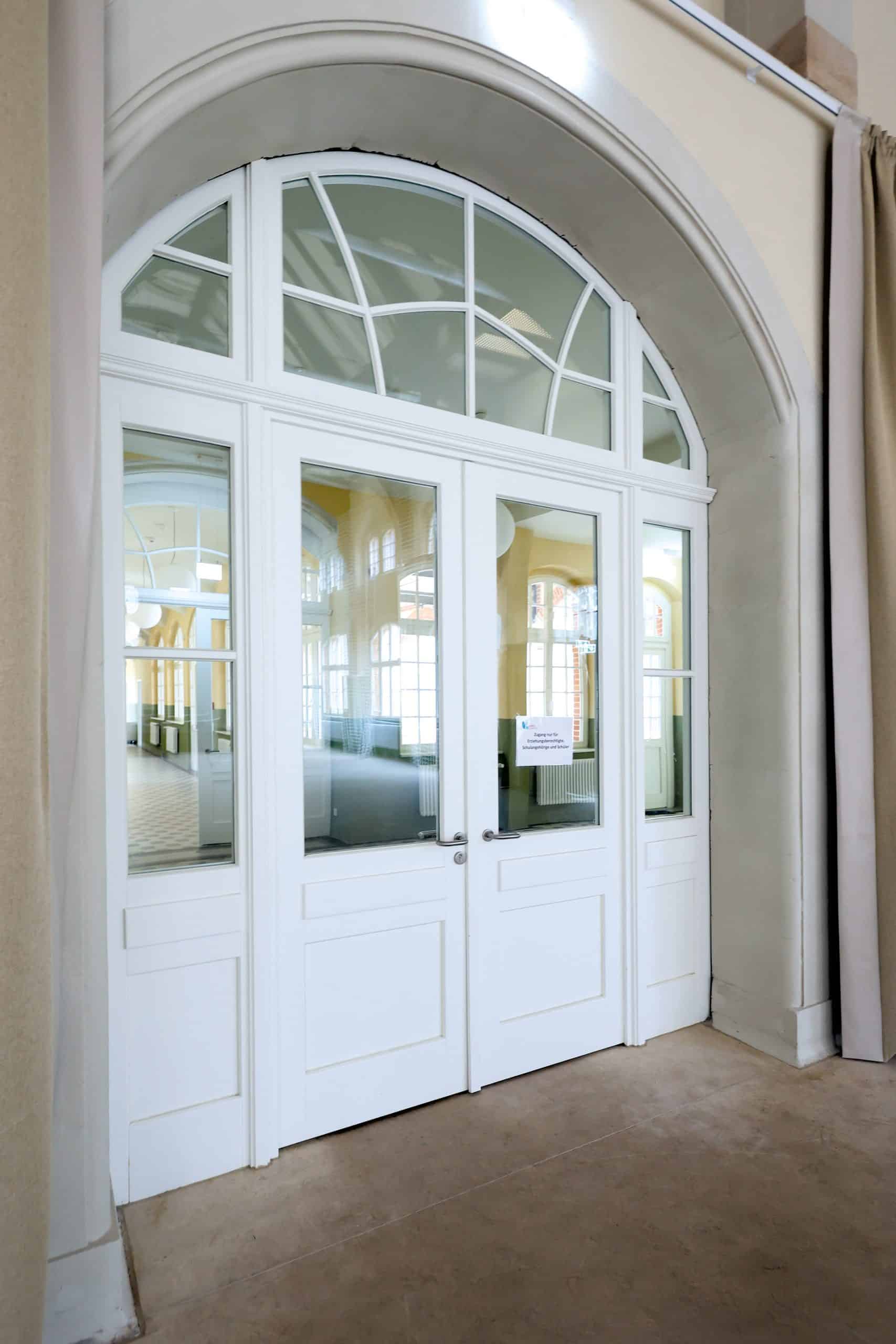 Ein Satz weißer Doppeltüren mit Glasfüllungen und einem gewölbten Oberlicht, die einen geräumigen, hellen Innenraum mit hohen Decken und großen Fenstern widerspiegeln. An einer der Türen ist ein kleines Schild angebracht, das von der Brandschutz Tischlerei Beelitz angefertigt wurde.