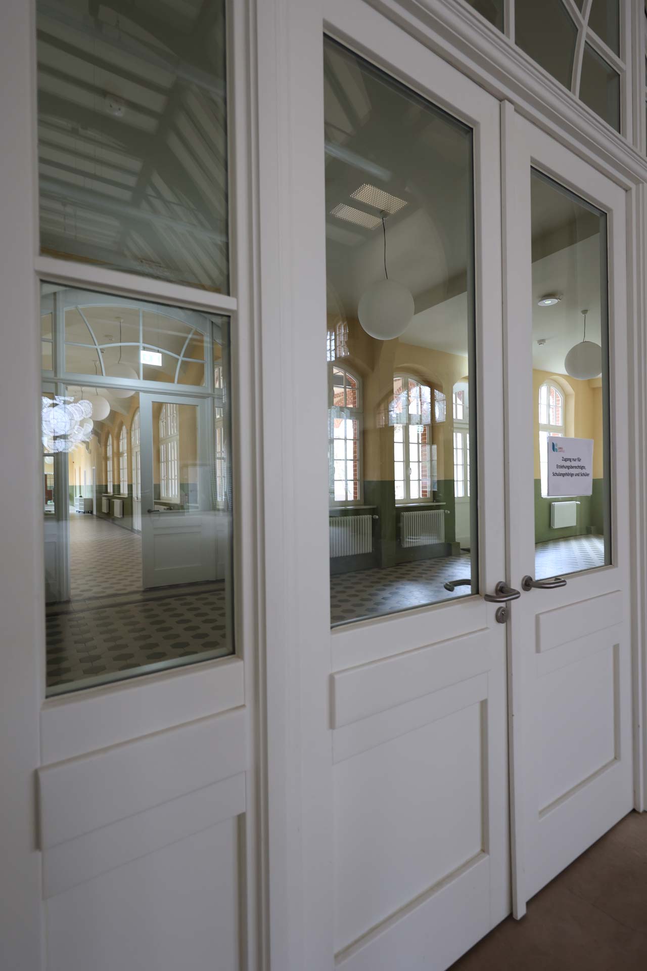 Weiße Doppeltüren mit Glasscheiben, möglicherweise eine Brandschutztür der Brandschutz Tischlerei Beelitz, geben den Blick frei auf einen großen, leeren, sonnendurchfluteten Raum mit hohen Bogenfenstern, runden Hängelampen und einem gemusterten Fliesenboden. An der rechten Tür ist ein kleines Schild angebracht.