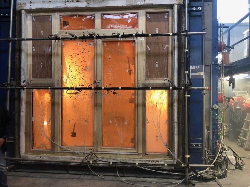Ein großes Brandschutzfenster wird in einem Labor einem Feuerwiderstandstest unterzogen. Flammen und intensives orangefarbenes Licht sind hinter dem Glas sichtbar, während Kabel und Sensoren der Brandschutz Tischlerei Beelitz den Fensterrahmen überwachen.