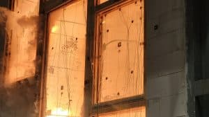 Große Fenster, möglicherweise Brandschutzfenster, sind starken Flammen und Hitze ausgesetzt, wobei das Glas zerspringt und Rauch aus der linken Seite aufsteigt - ein Hinweis darauf, dass ein Brand oder ein Feuerwiderstandstest der Brandschutz Tischlerei Beelitz im Gange ist.