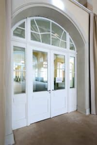 Ein Satz weißer Doppeltüren mit Glasfüllungen und einem gewölbten Oberlicht, die einen geräumigen, hellen Innenraum mit hohen Decken und großen Fenstern widerspiegeln. An einer der Türen ist ein kleines Schild angebracht, das von der Brandschutz Tischlerei Beelitz angefertigt wurde.