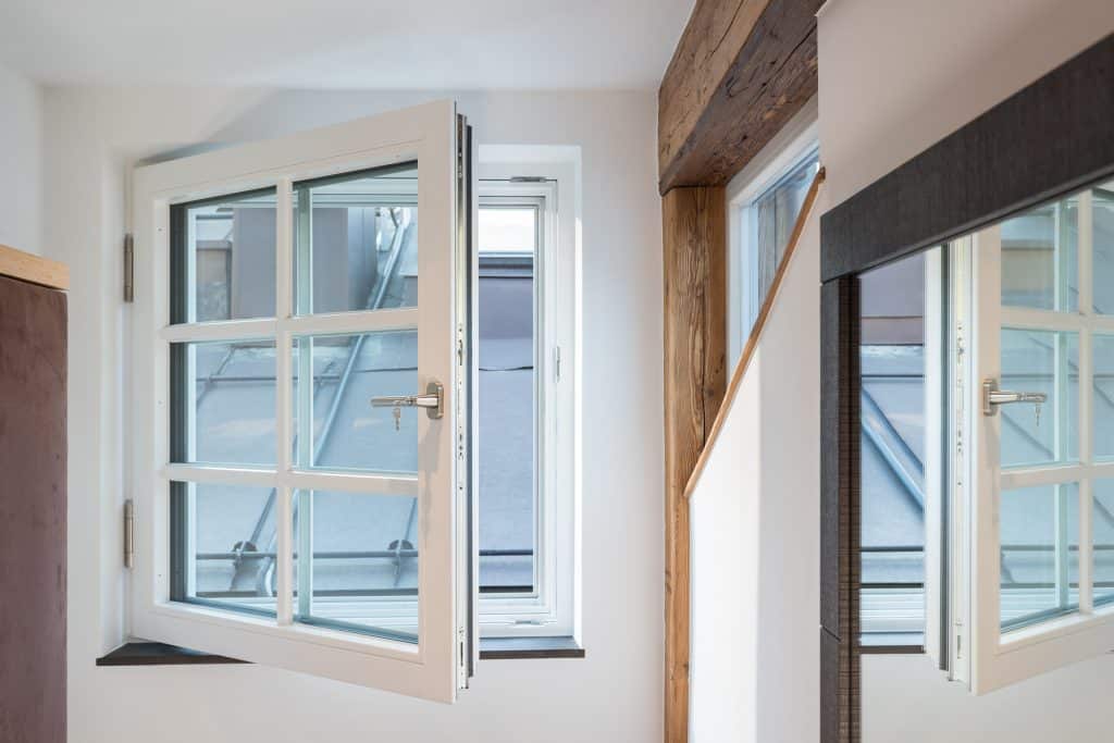 weiß gerahmtes Brandschutzfenster Ein weiß gerahmtes Brandschutzfenster ist geöffnet und gibt den Blick auf ein graues Dach frei. Der Raum hat weiße Wände, freiliegende Holzbalken und einen großen Wandspiegel, der einen Teil des Fensters und des Rauminneren reflektiert.