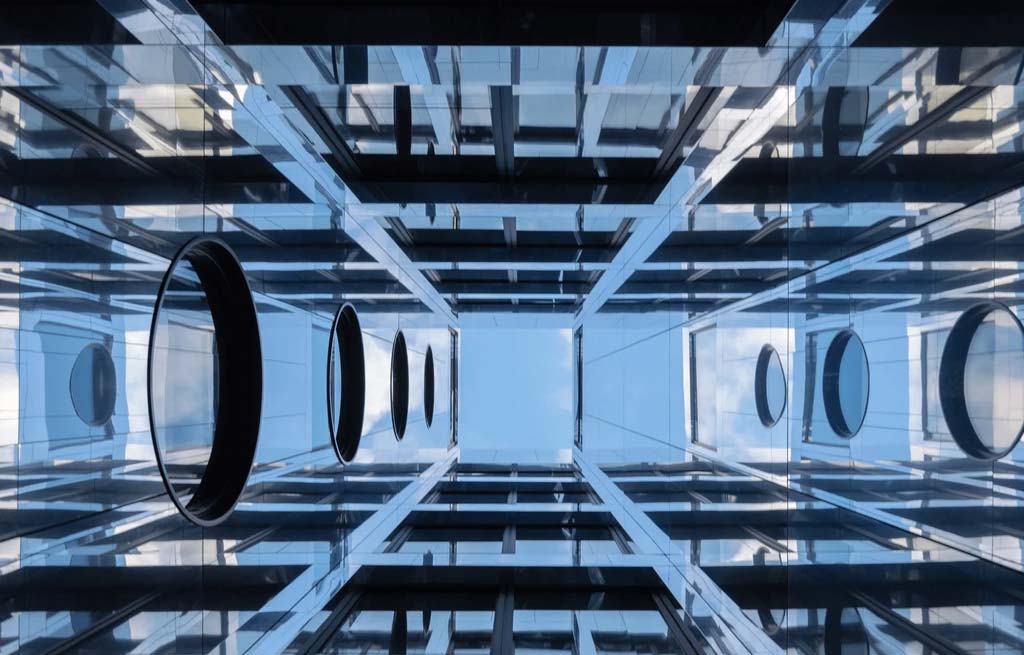 Blick auf ein modernes gläsernes Atrium mit geometrischen Linien, Spiegelungen und kreisförmigen schwarzen Ringen, die von der Decke vor einem blauen Himmel abgehängt sind, mit Brandschutzfenstern für mehr Sicherheit von der Brandschutz Tischlerei Beelitz.