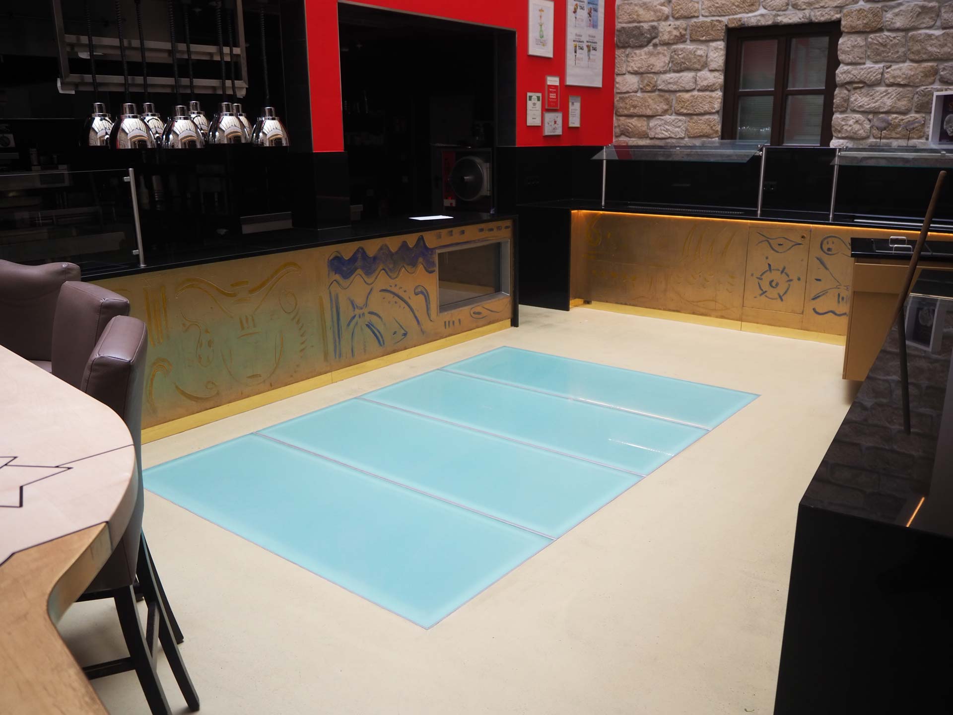 Eine moderne Küche mit einer dekorativen gelb-schwarzen Theke, braunen Stühlen und einer großen Milchglasscheibe, die in den hellen Boden eingelassen ist. Im Hintergrund sind eine freiliegende Steinwand, ein Fenster und eine schlichte Brandschutztür zu sehen.