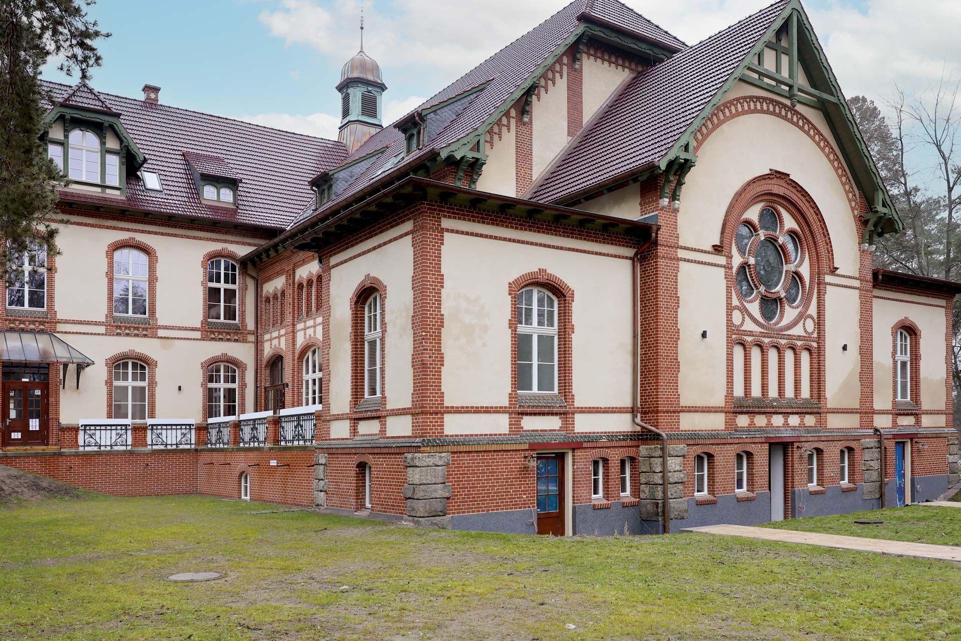 Historisches Backsteingebäude mit Rundbogenfenstern, einem runden Buntglasfenster, steilen Giebeldächern und einem kleinen Turm mit Brandschutzfenster und Brandschutztür der Brandschutz Tischlerei Beelitz, umgeben von Gras und Bäumen unter einem teilweise bewölkten Himmel.