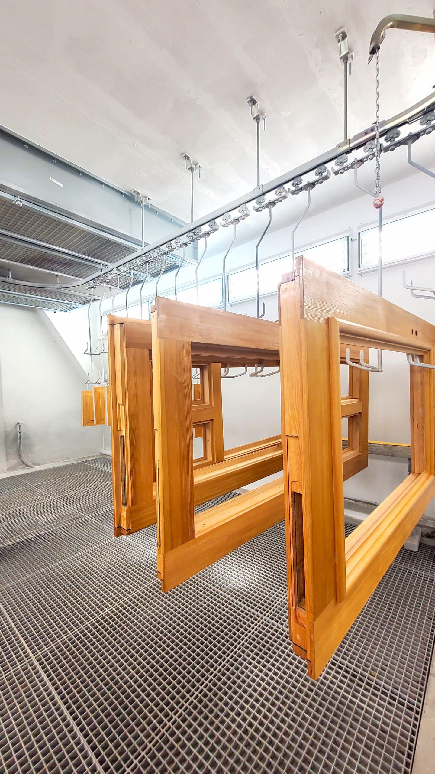 Holzrahmen für Brandschutztür oder Brandschutzfenster der Brandschutz Tischlerei Beelitz, hängen an einem Metallschienensystem in einer Industriehalle mit heller Beleuchtung und einem darunter liegenden Metallgitterboden.