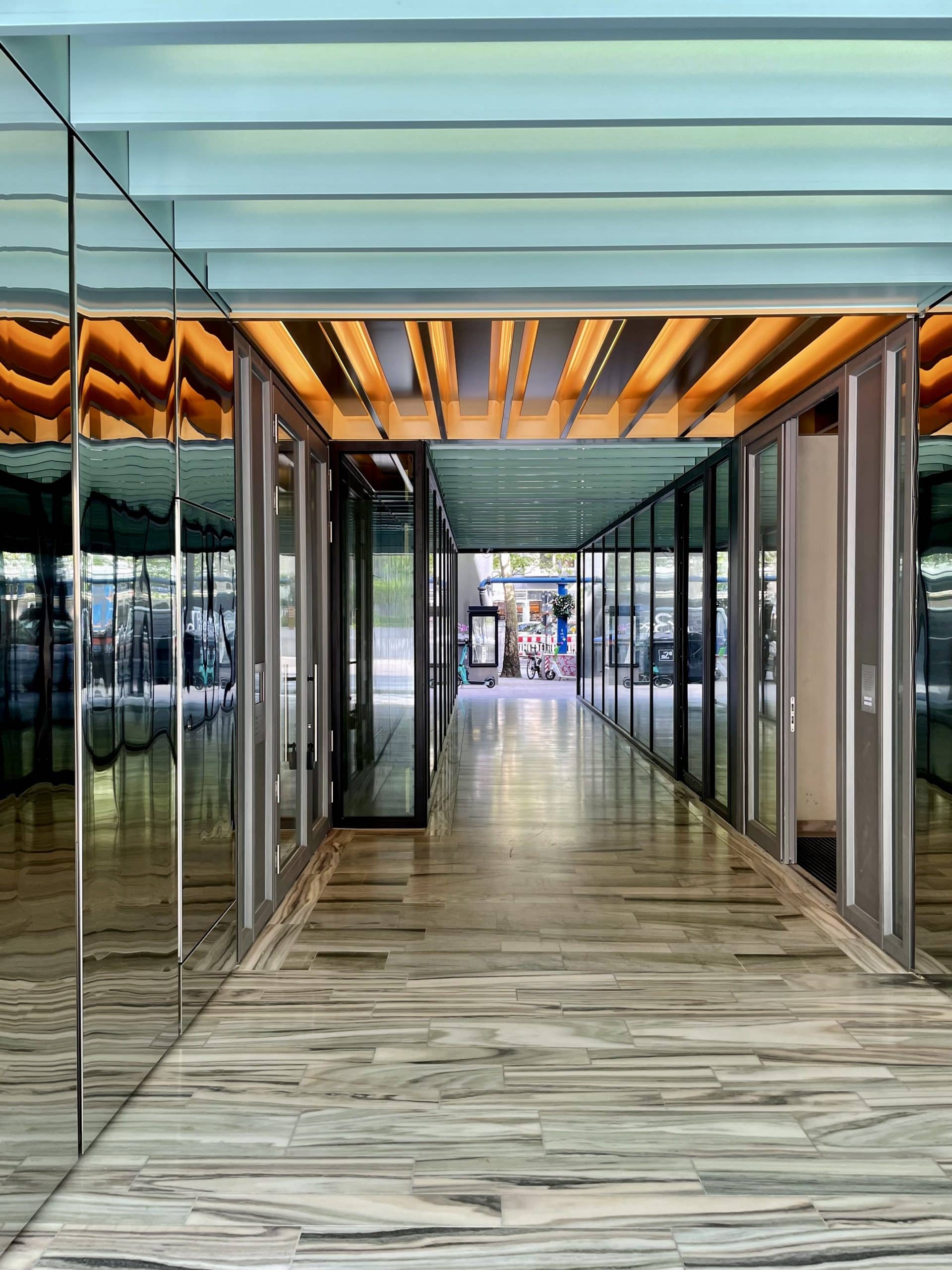 Ein moderner Flur mit Glaswänden, verspiegelten Paneelen und gestreiften Holzböden verfügt über eine Brandschutztür und eine Decke aus abwechselnd blau und orange beleuchteten Paneelen, die zu einem Außenbereich mit Bäumen in der Ferne führt.