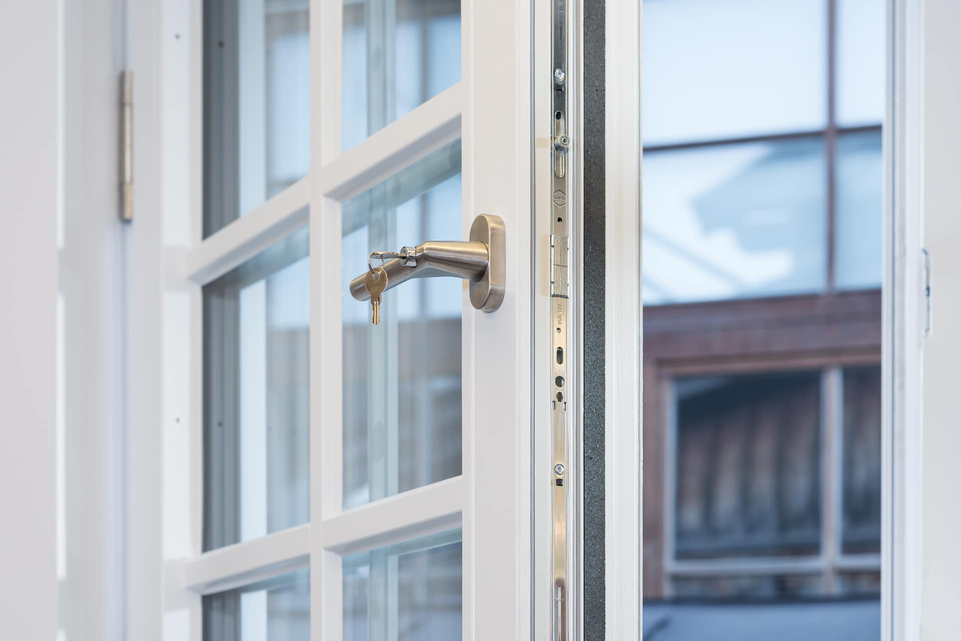 Nahaufnahme eines weißen Brandschutzfensters mit Glasscheiben, leicht geöffnet, mit Metallgriff und Schlüssel im Schloss; außerhalb des Fensters ist ein Gebäude zu sehen.