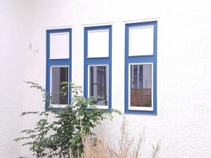 Drei schmale rechteckige Brandschutzfenster mit blauen Rahmen sind in eine weiß strukturierte Wand eingelassen. Vor den Fenstern wächst ein grüner, belaubter Strauch, und am unteren Rand des Bildes ist trockenes Gras zu sehen.