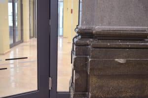 Nahaufnahme einer großen Steinsäule neben einer Brandschutztür in einem Hausflur mit beigem Kachelboden und gelben Wänden. Der Bereich hinter der Tür, die von der Brandschutz Tischlerei Beelitz hergestellt wurde, ist leer und hell erleuchtet.