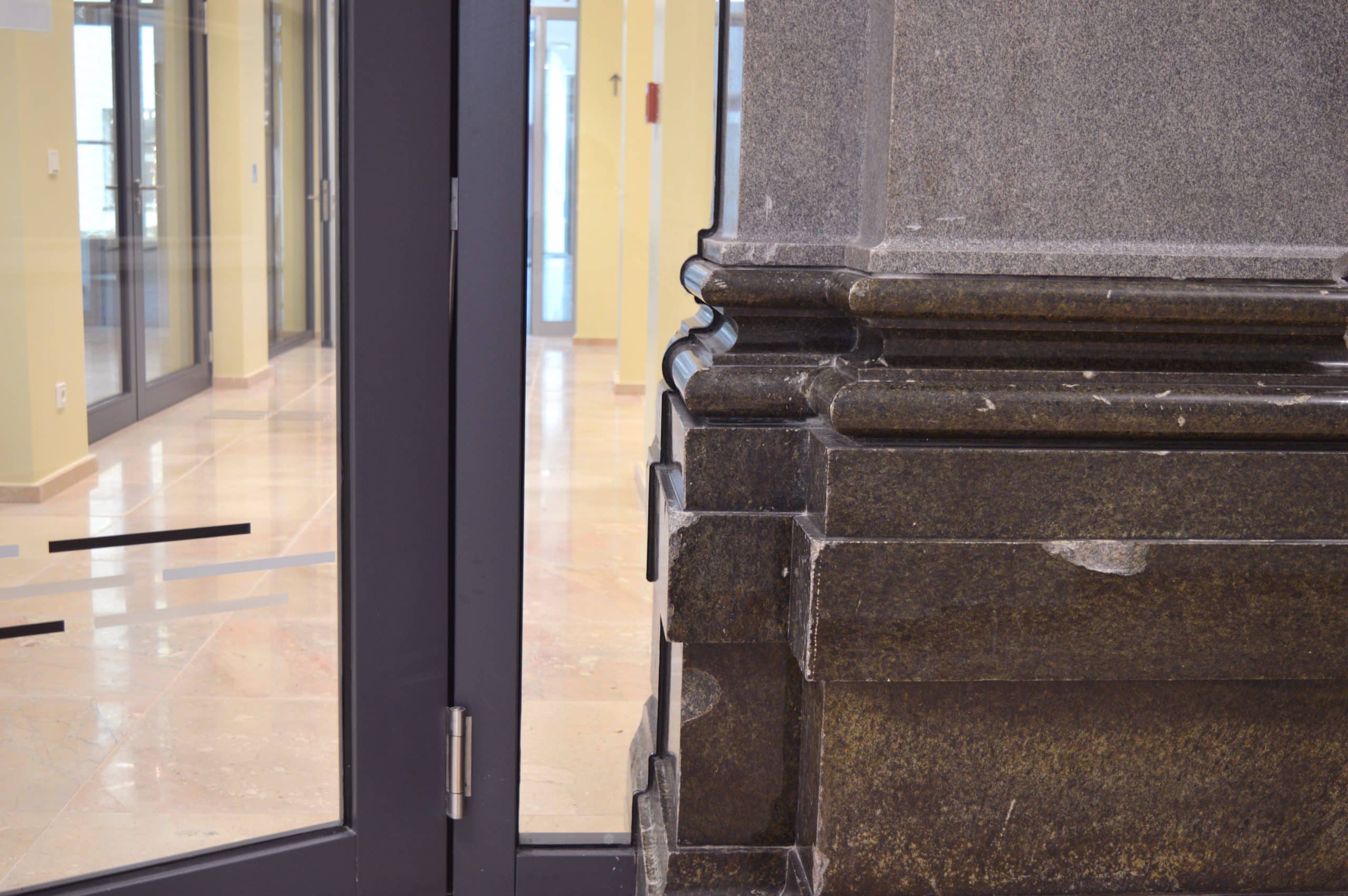 Nahaufnahme einer großen Steinsäule neben einer Brandschutztür in einem Hausflur mit beigem Kachelboden und gelben Wänden. Der Bereich hinter der Tür, die von der Brandschutz Tischlerei Beelitz hergestellt wurde, ist leer und hell erleuchtet.