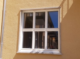 Ein weiß gerahmtes Brandschutzfenster mit vier Glasscheiben ist in eine beige strukturierte Wand eingelassen und spiegelt ein Gebäude und den blauen Himmel wider. Oberhalb des Fensters sind die Schatten eines Geländers zu sehen.